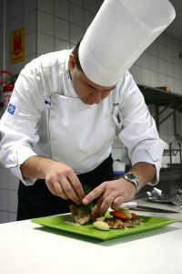 Chef in a white uniform and hat arranging food on a green plate in a kitchen. - International Hotel School