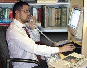 A man in a white shirt and tie using a telephone while working on a computer at a desk. - International Hotel School