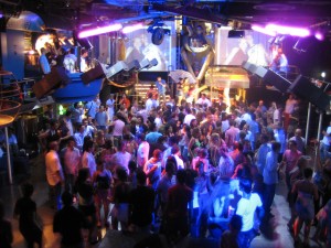 A lively crowd dancing under colorful lights in a nightclub setting with a DJ booth. - International Hotel School
