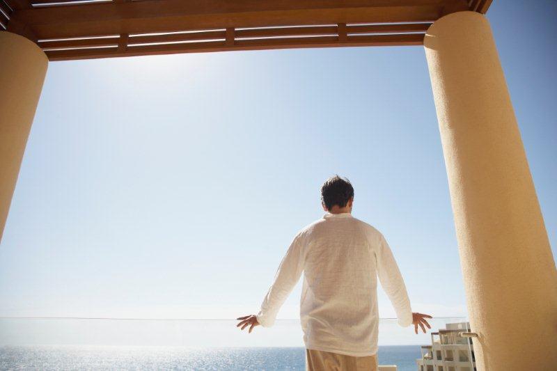 A man in a white shirt standing on a balcony, gazing at the ocean under a clear sky. - International Hotel School