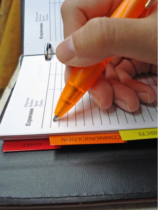 A hand holding an orange pen writing in a notebook with organized tabs for different subjects. - International Hotel School