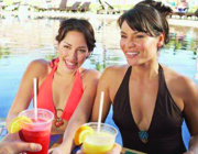 Two women in swimsuits holding colorful drinks with lemon slices while sitting by a pool. - International Hotel School