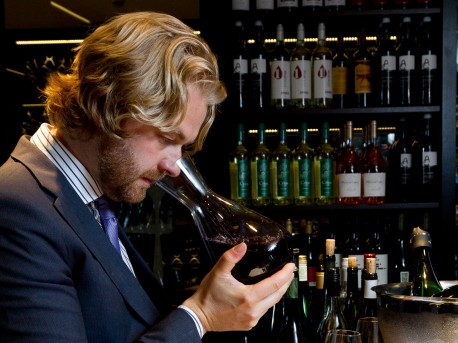 Sommelier examining a glass of red wine while wearing a suit in a bar setting. - International Hotel School