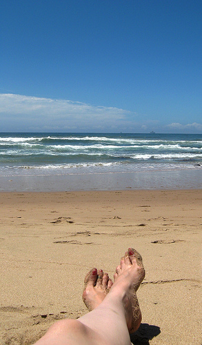 Bare feet resting on sandy beach with ocean waves in the background under a clear blue sky. - International Hotel School