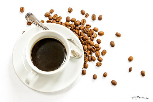 A white cup filled with black coffee placed on a saucer, surrounded by coffee beans. - International Hotel School