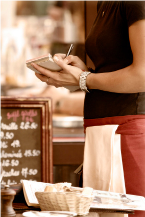 A waitress in a black shirt and apron writing down an order on a notepad at a restaurant. - International Hotel School
