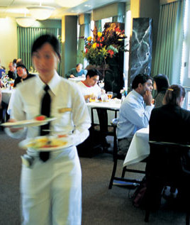 Waitstaff in uniform carrying plates of food in a dining area with patrons seated at tables. - International Hotel School