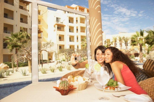 Two women smiling and taking a selfie at a restaurant table with drinks and food. - International Hotel School