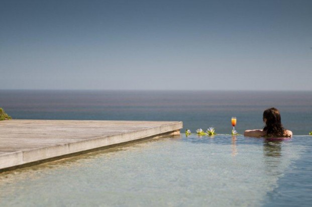 Individual sitting at the edge of a pool holding a drink, overlooking a calm body of water. - International Hotel School