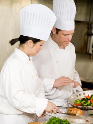 Two chefs in white uniforms and tall hats working together in a kitchen, preparing vegetables and meat. - International Hotel School