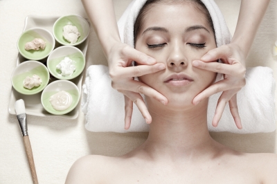 A woman undergoing a facial treatment with hands applying pressure on her face and various creams in bowls nearby. - International Hotel School