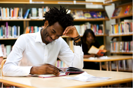A male student with dreadlocks reading a book at a table in a library. - International Hotel School