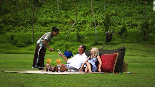 A couple relaxing outdoors while being served refreshments by a staff member in a tropical setting. - International Hotel School