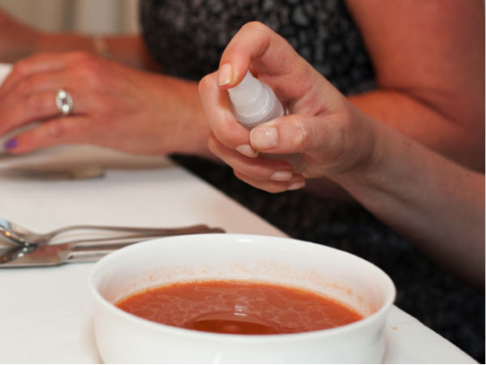 A hand holding a spray bottle above a bowl of soup on a dining table. - International Hotel School
