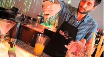 Bartender in a striped shirt pouring orange juice into a glass at a bar. - International Hotel School