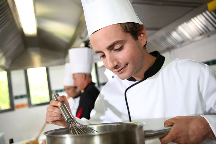 Chef in a white uniform and hat mixing ingredients in a stainless steel pot in a kitchen. - International Hotel School