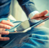 Individual interacting with a tablet device while seated, wearing a blue jacket and jeans. - International Hotel School