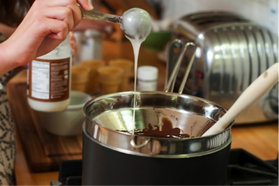 A person using a measuring spoon to add coconut oil into a pot on a stove in a kitchen. - International Hotel School
