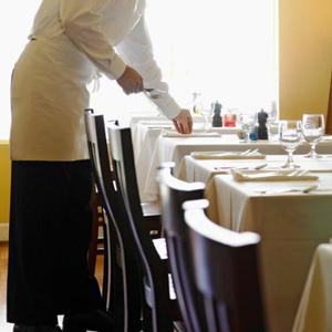A waiter adjusting table settings in a restaurant with white tablecloths and glassware. - International Hotel School