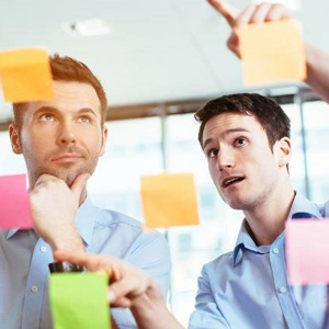 Two men in light blue shirts discussing ideas while pointing at colorful sticky notes on a glass wall. - International Hotel School