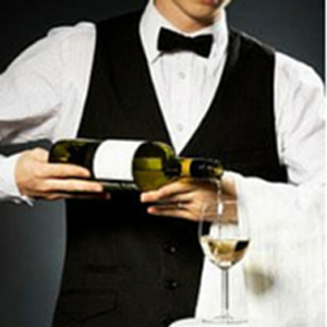 A server in a black vest and bow tie pouring white wine from a bottle into a glass. - International Hotel School