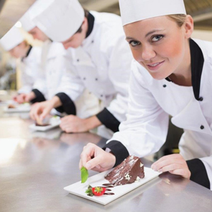 A female chef garnishing a dessert plate with chocolate and a strawberry in a kitchen setting. - International Hotel School