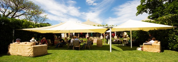 Large outdoor tent with seating area and people gathered underneath in a garden setting. - International Hotel School