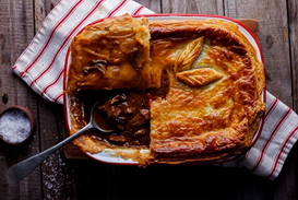 A mushroom pie with a golden-brown crust and a serving spoon resting beside it on a wooden surface. - International Hotel School