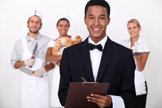 A male culinary student in a suit holding a clipboard with three peers in the background wearing chef uniforms. - International Hotel School