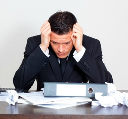 A businessman in a suit with his hands on his head, looking stressed at a desk filled with papers and a binder. - International Hotel School