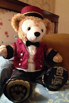 A teddy bear dressed in a red hotel uniform with a bow tie and black pants, sitting on a bed. - International Hotel School