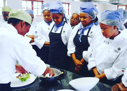 Culinary students observing a chef preparing a dish in a kitchen setting. - International Hotel School