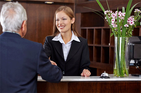 A hotel receptionist in a black suit greeting a guest at the front desk with a smile. - International Hotel School