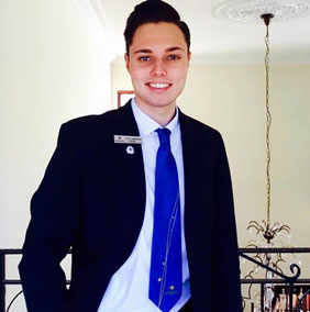 A hospitality student wearing a black suit and blue tie, standing indoors with a confident smile. - International Hotel School