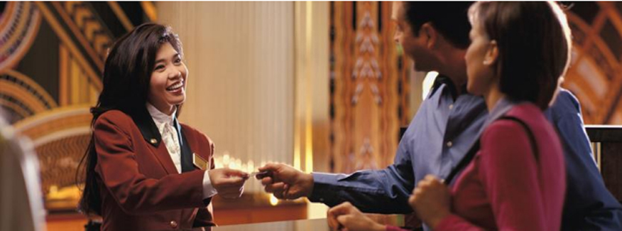 Hotel receptionist in a red blazer handing a key card to guests at a reception desk. - International Hotel School