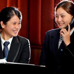 Two women in formal attire, one smiling and the other speaking on the phone, in a professional setting. - International Hotel School