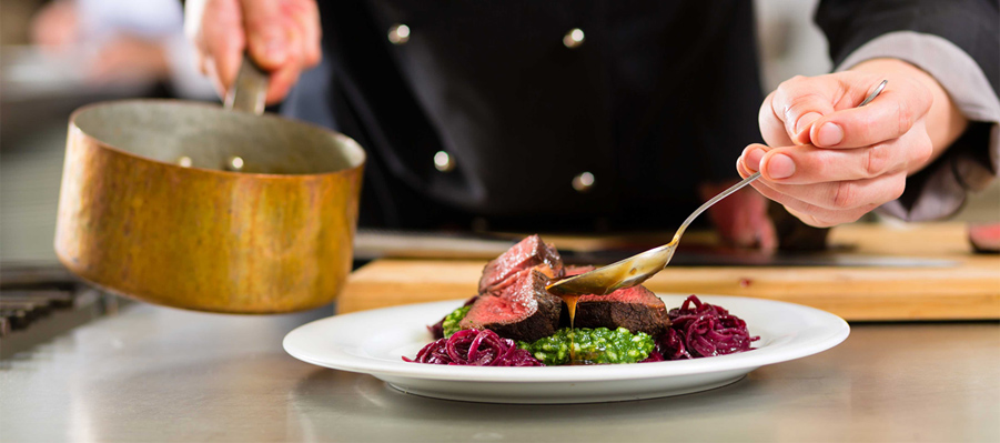 A chef plating a steak with sauce and garnishes in a professional kitchen setting. - International Hotel School