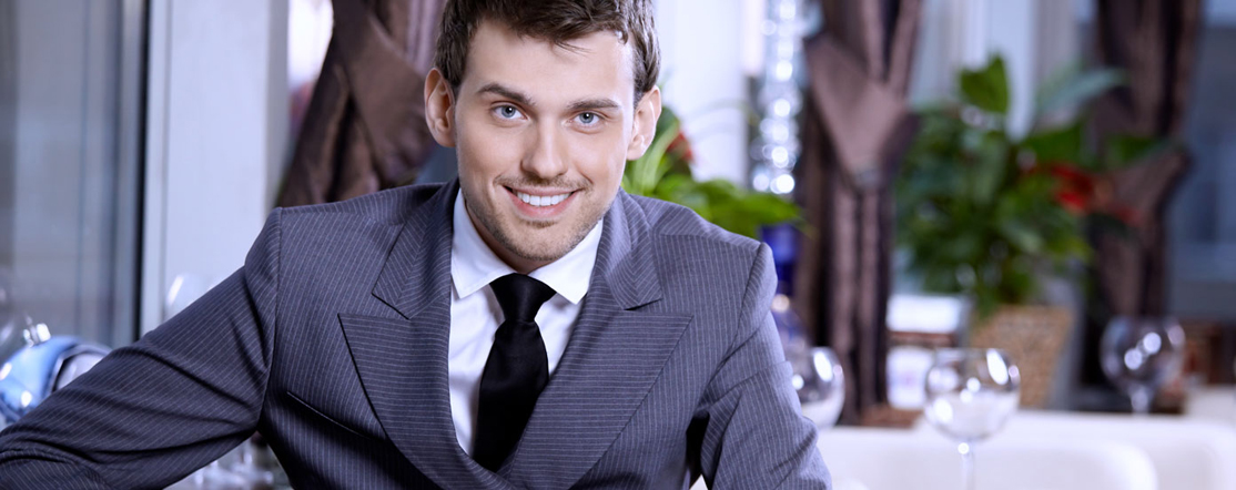 A man in a gray suit and black tie smiling while seated at a table with glassware. - International Hotel School