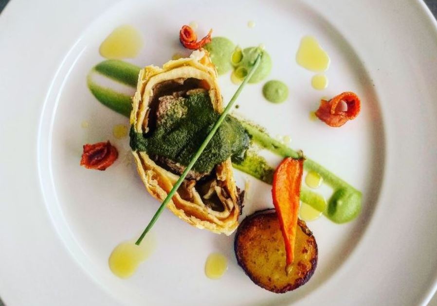 Lamb Wellington served with green sauce and garnished with vegetables on a white plate. - International Hotel School
