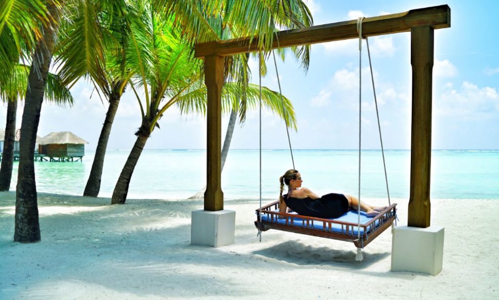 A woman relaxing on a swing bed under palm trees on a beach in the Maldives. - International Hotel School