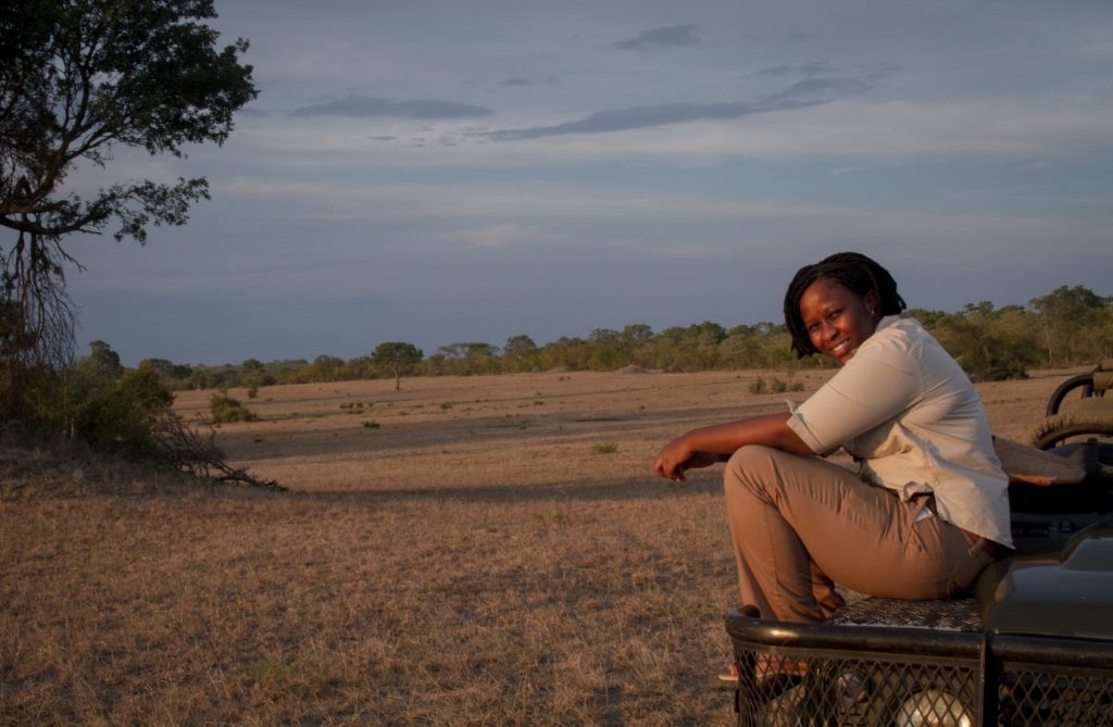 A woman sitting on the edge of a safari vehicle in a natural landscape during sunset. - International Hotel School