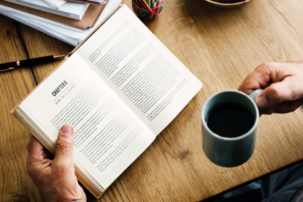 An open book displaying text on a wooden table with a person's hand holding a coffee mug. - International Hotel School