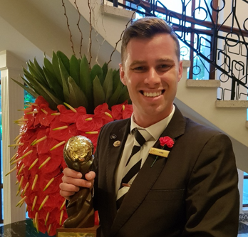 Man in a black suit holding a trophy, standing in front of a decorative plant. - International Hotel School