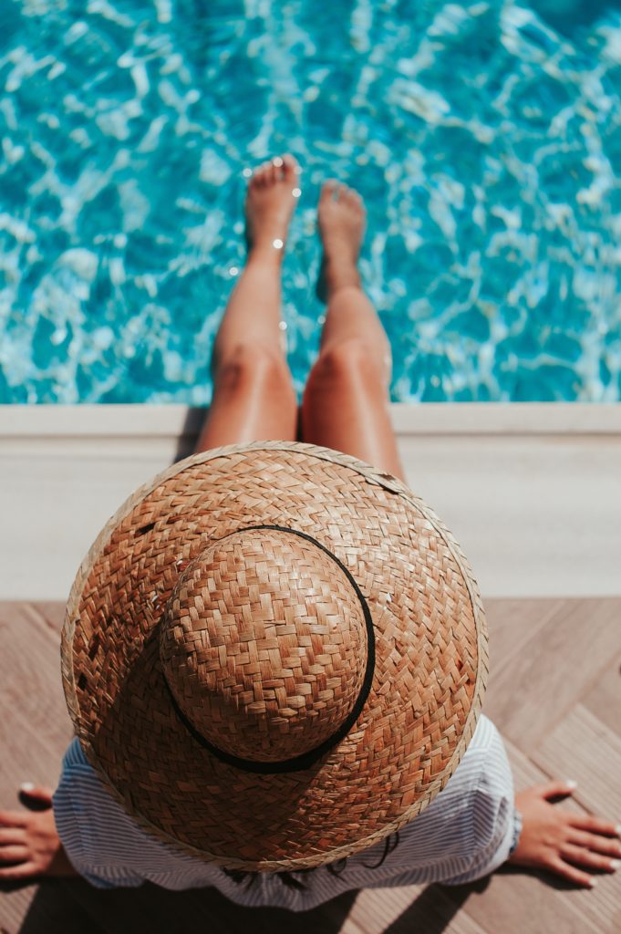 Individual wearing a straw hat sitting by the edge of a swimming pool with feet in the water. - International Hotel School