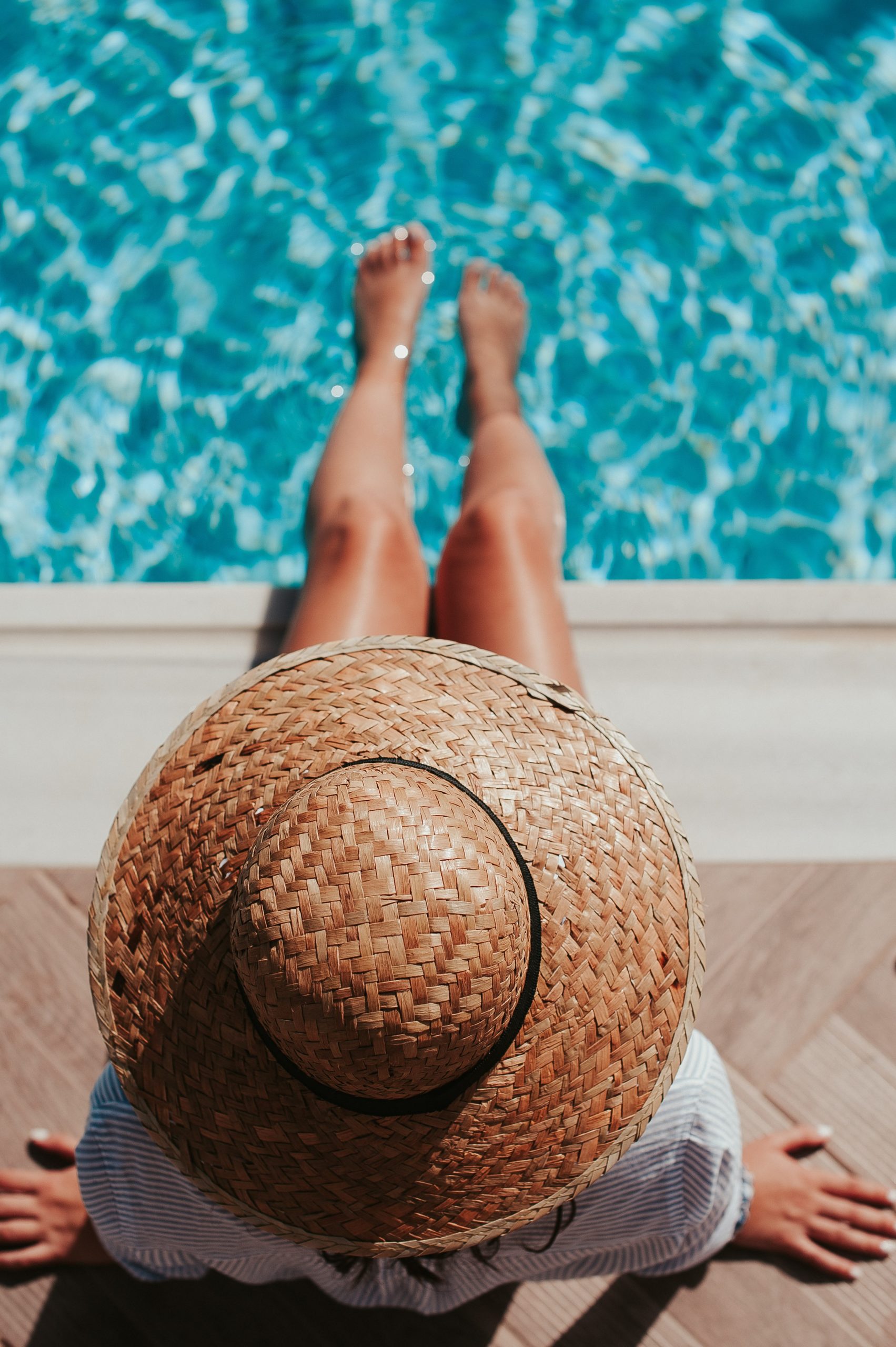 Individual wearing a straw hat sitting by the edge of a swimming pool with feet in the water. - International Hotel School