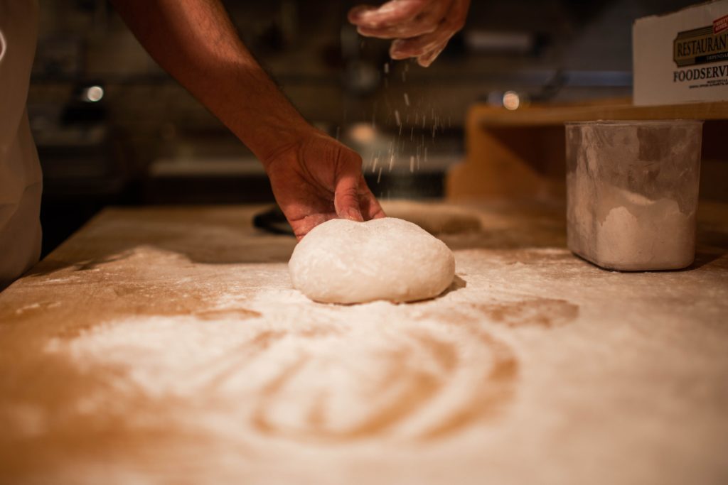 A hand sprinkling flour over a round piece of dough on a wooden surface. - International Hotel School