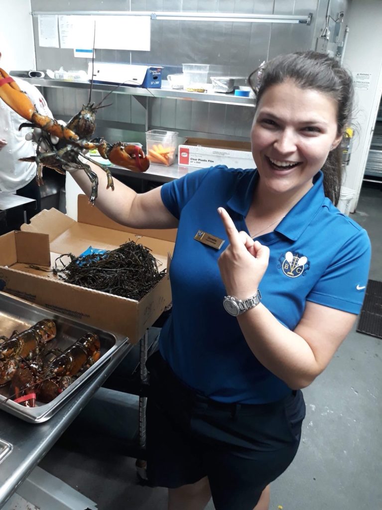 A woman in a blue shirt holding a large lobster while smiling in a kitchen environment. - International Hotel School