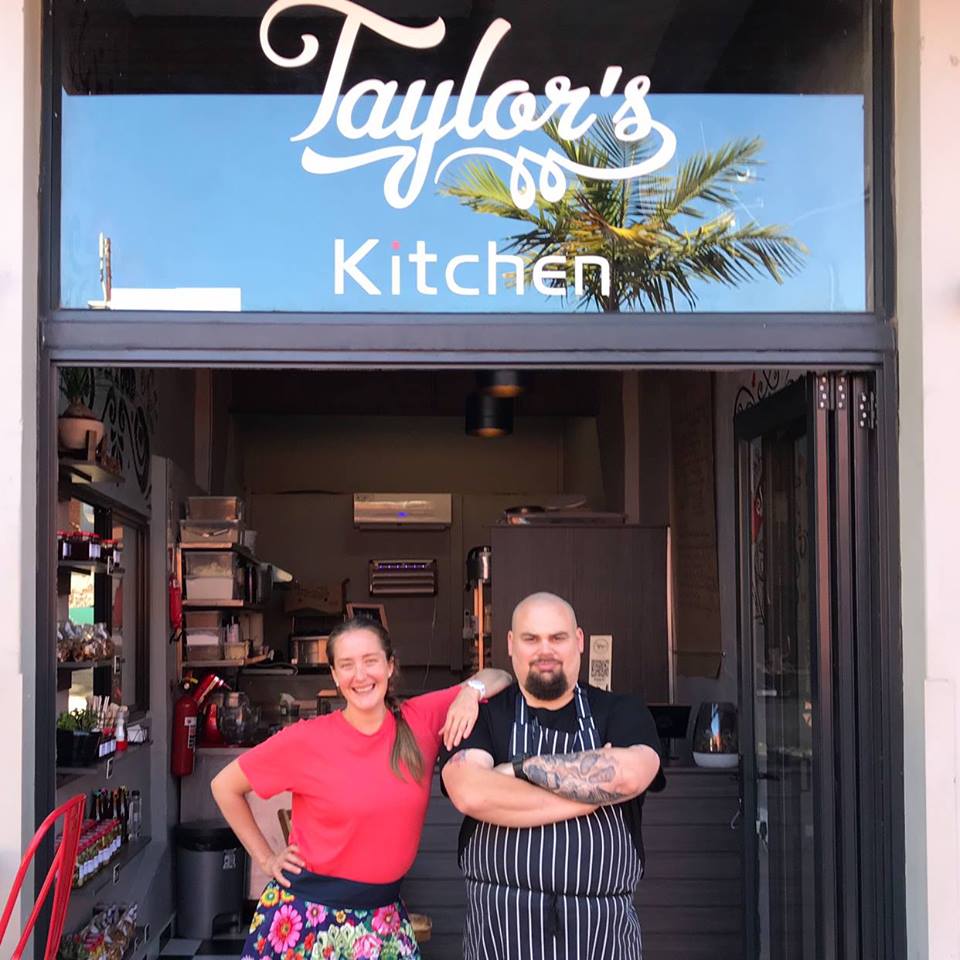 Two staff members standing at the entrance of Taylor's Kitchen, a restaurant with a sign above. - International Hotel School