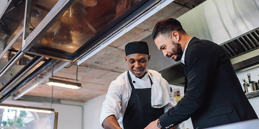 A chef in a white uniform and a businessman discussing food preparation in a modern kitchen. - International Hotel School