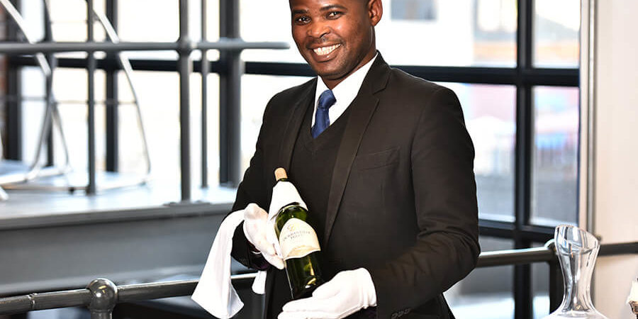 Sommelier in formal attire presenting a bottle of wine with a white cloth in a professional setting. - International Hotel School